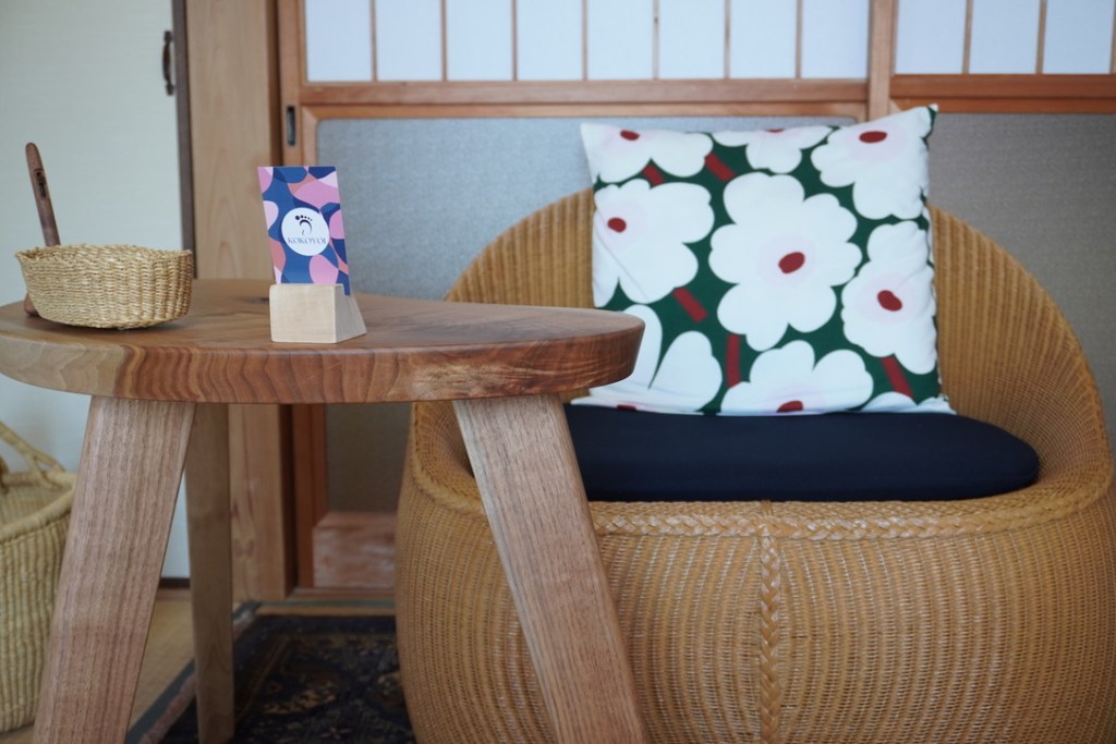 A cozy seating area featuring a round woven chair with a decorative floral cushion, a wooden table with a small card holder displaying KOKOYOI branding, and a woven basket beside the table.
